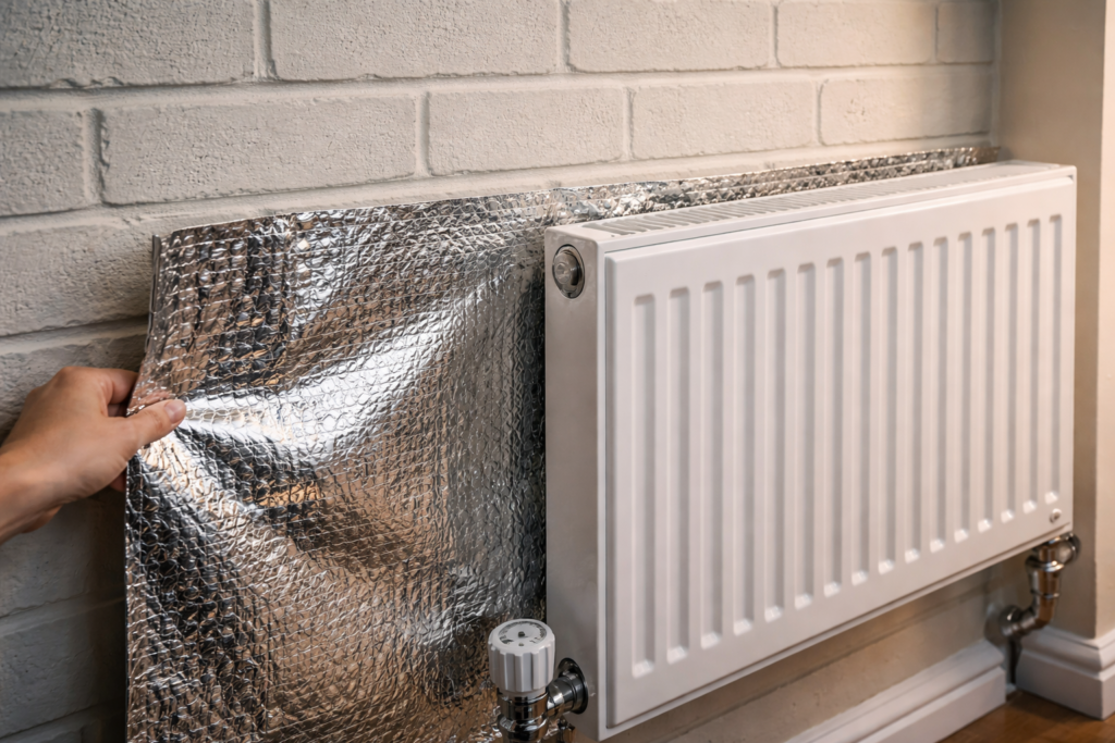 Silver reflective radiator foil being installed behind a white panel radiator on an external brick wall in a UK home