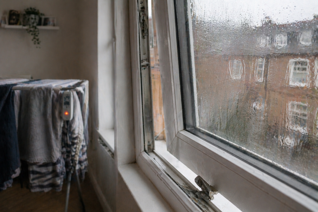 Light condensation on single glazed window in small UK room with folding heated airer loaded with clothes visible in background and window slightly ajar